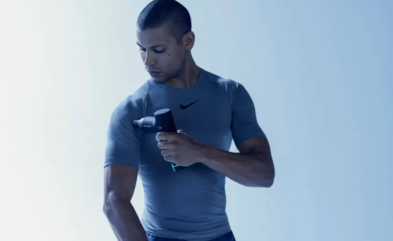 Muscular man in Nike shirt using Hypervolt GO massage gun on his arm, blue background