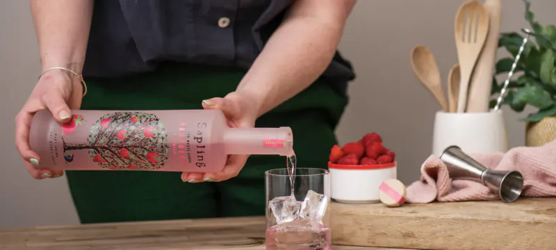 Woman pouring pink PINKTREE gin from bottle into ice-filled glass with strawberries on wooden surface