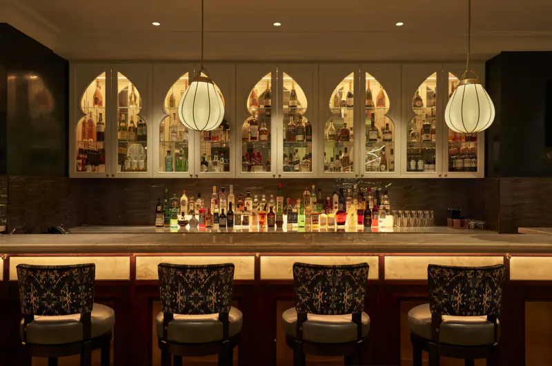 Elegant hotel bar with arched glass cabinets of colorful liquor bottles, pendant lights, and upholstered stools at marble counter.