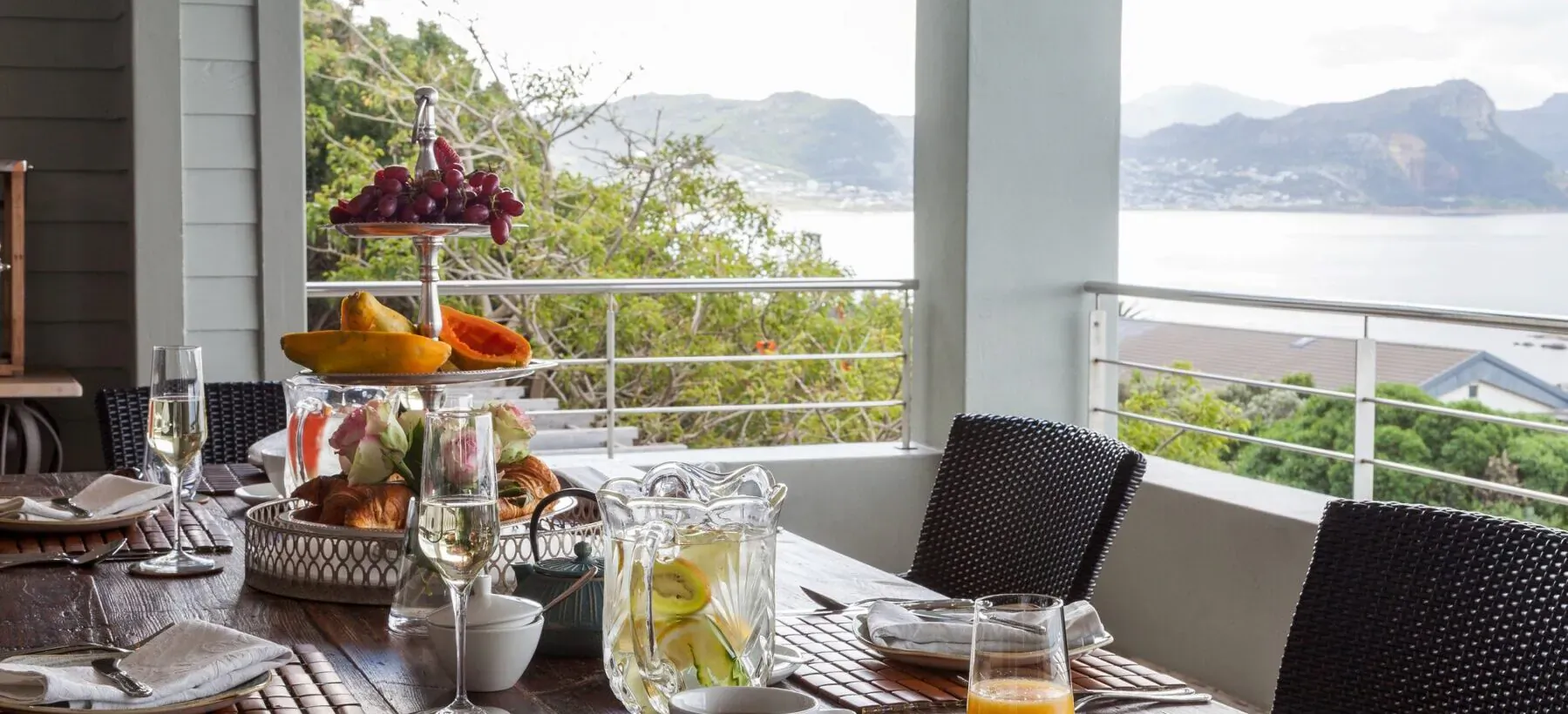 Elegant outdoor table at Tintswalo Boulders Hotel with fruit platters, drinks, overlooking Simon's Town sea and mountains.