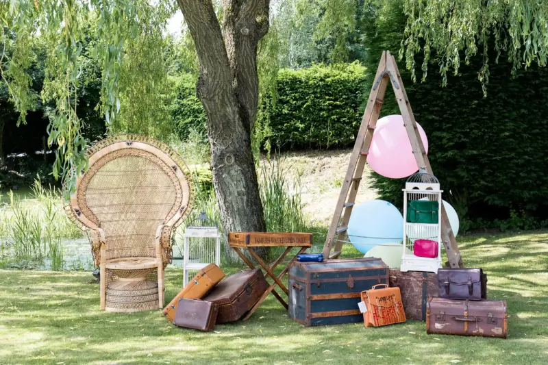 Rustic garden photo booth setup with rattan chair, vintage leather luggage, balloons, ladder, and lanterns under trees.