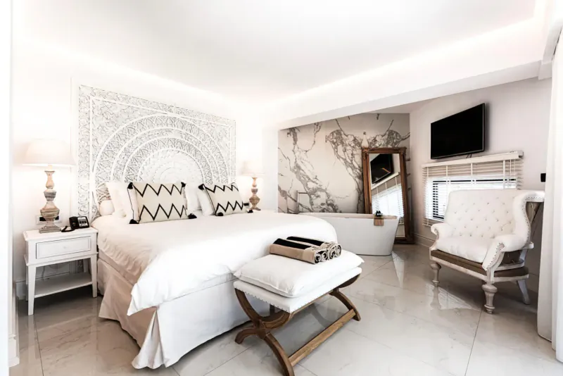 Elegant white bedroom at Abaton Island Resort & Spa with king bed, marble bathtub, mandala headboard, and armchair.