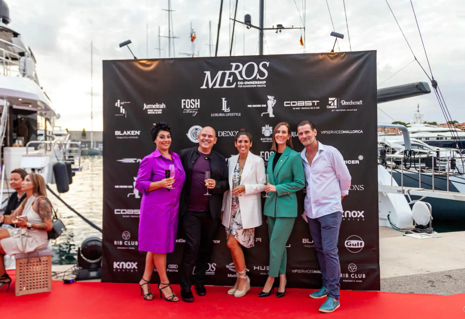 Group of 6 elegantly dressed people posing on red carpet at Marinos event backdrop in marina with yachts, Balearic Islands.