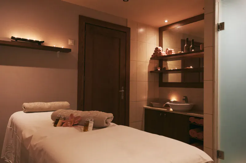 Luxurious spa room with white massage table topped with towels and hibiscus flower, lit shelves, sink, and frosted glass shower door.