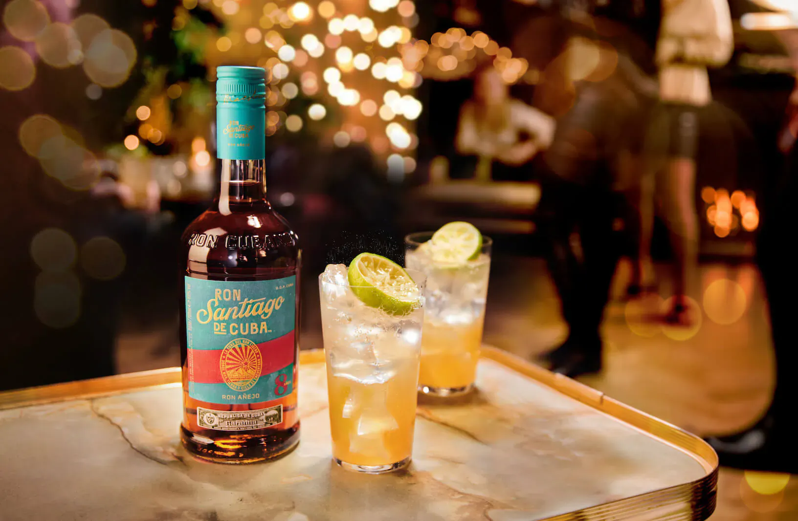 Ron Salvaje Cuba rum bottle and two highball glasses with lime wedges on marble table in festive bar setting