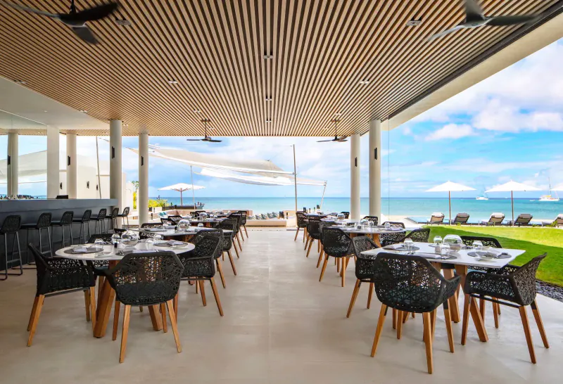 Open-air beachfront dining at Silversands Grenada with wicker tables, chairs, bar, umbrellas, and ocean view.
