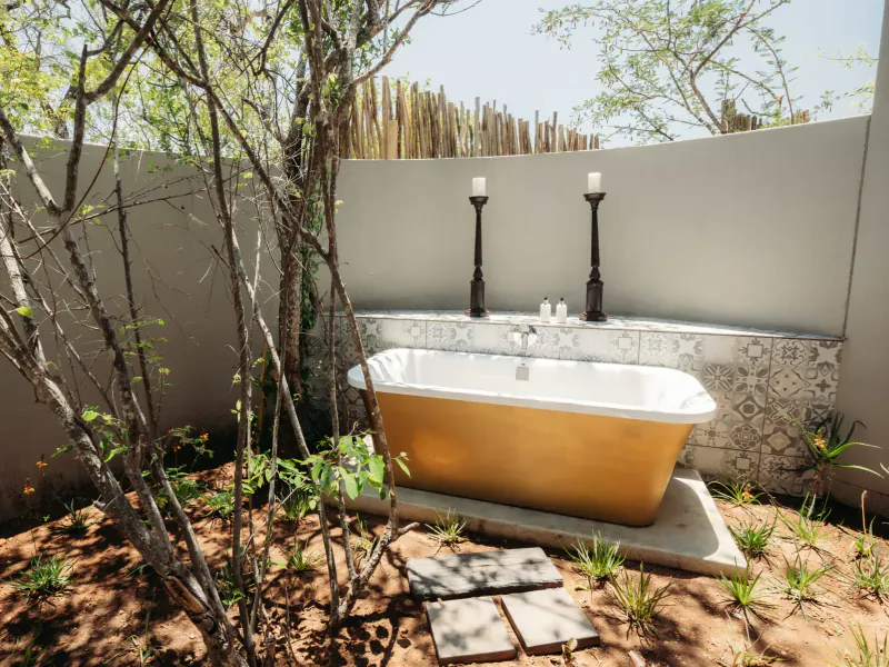 Gold bathtub in outdoor safari lodge setting with trees, plants, tiled wall, and lit candelabras