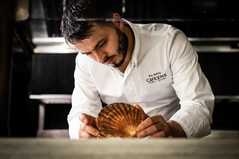 Chef in No. Fifty Cheyne uniform closely examining a large scallop shell in professional kitchen