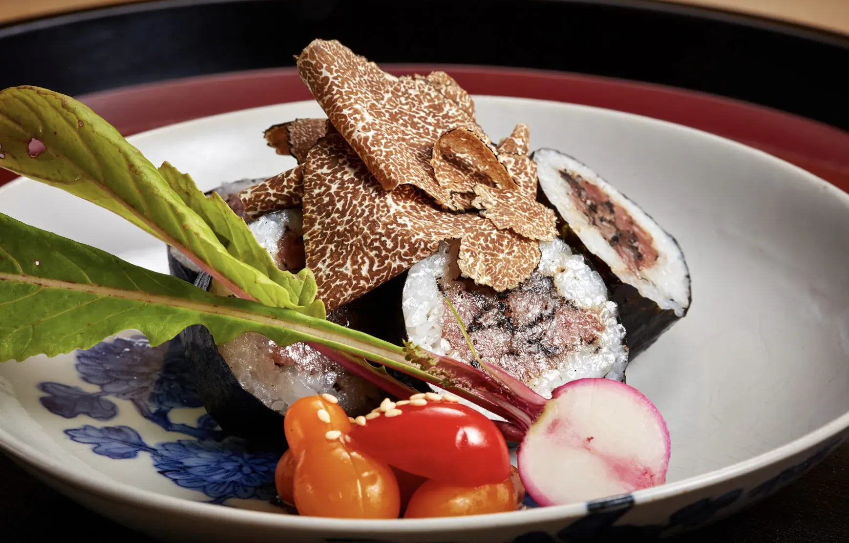 Kaiseki sushi roll topped with crispy truffles, garnished with red leaf, radish, cherry tomatoes in blue floral bowl