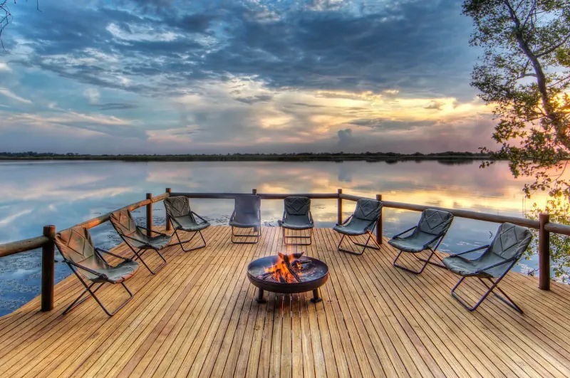 Sunset view of wooden deck on lake at Xugana Island Lodge with fire pit and canvas chairs