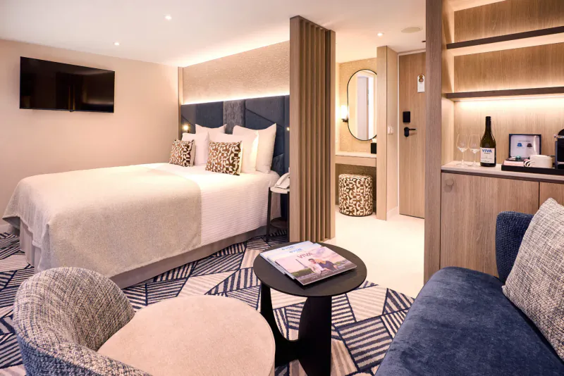 Spacious modern cruise ship suite with king bed, blue sofa, coffee station, wine bottle, and TV.