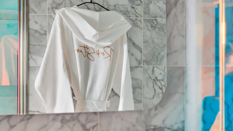 White hooded bathrobe with 'MVJ' logo hanging on hook against marble bathroom wall, Virgin Cruises.