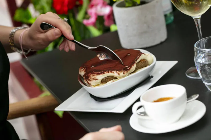 Hand with spoon scooping tiramisu from white bowl on square plate, beside coffee cup, wine glass, water, and pink flowers on table.