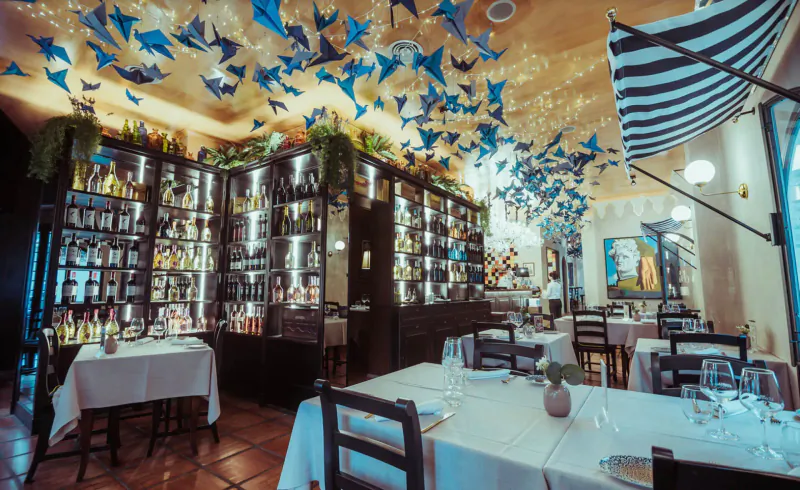 Elegant Italian restaurant interior with blue paper birds hanging from ceiling, wooden shelves of liquor bottles, and white-clothed dining tables.
