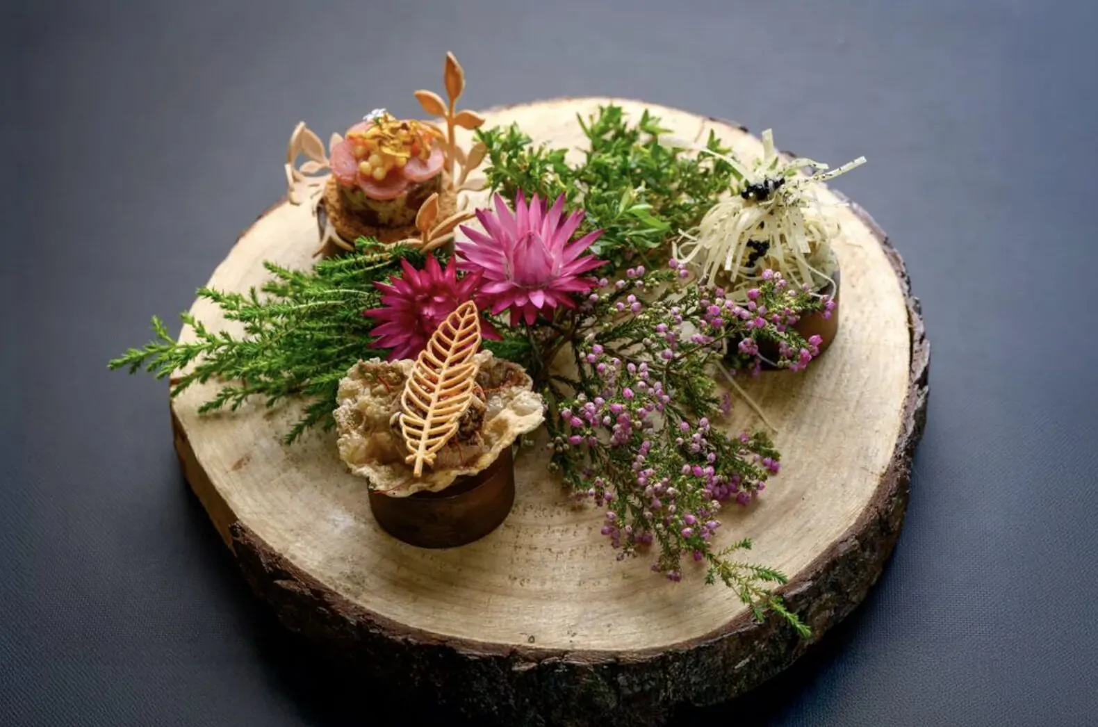 Vibrant floral arrangement of pink daisies, orange flowers, ferns, and mushrooms on a wooden slice by Forage Chef Gregory Henderson