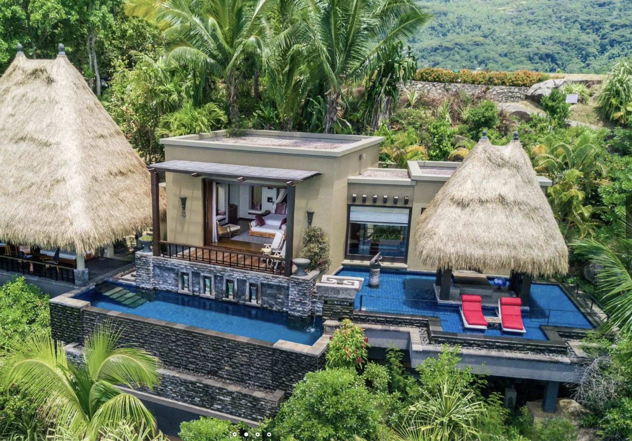 Luxury thatched villa with infinity pool, red lounge chairs, and open bedroom, perched in lush tropical jungle with mountains.