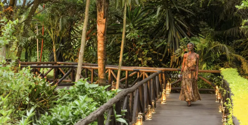 Woman in flowing dress walks lantern-lit wooden bridge through lush tropical safari gardens at Fairmont Mara Safari Club