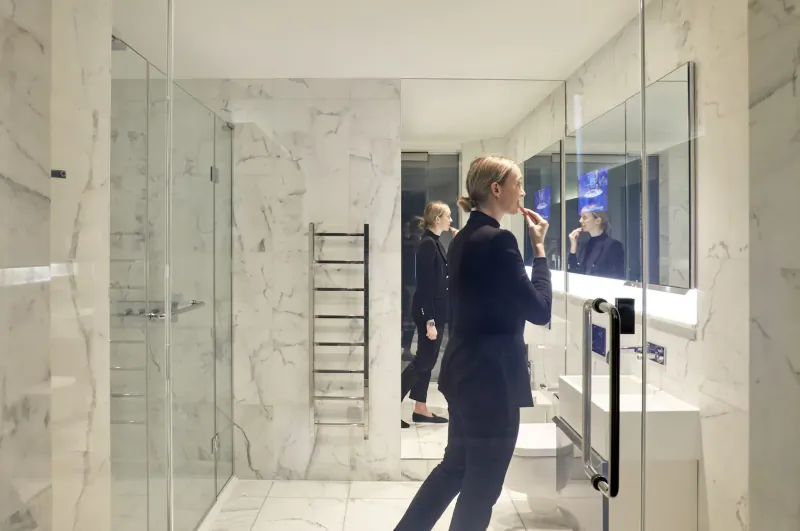 Woman in black suit applying makeup at mirror in luxurious white marble bathroom at Eccleston Square Hotel London