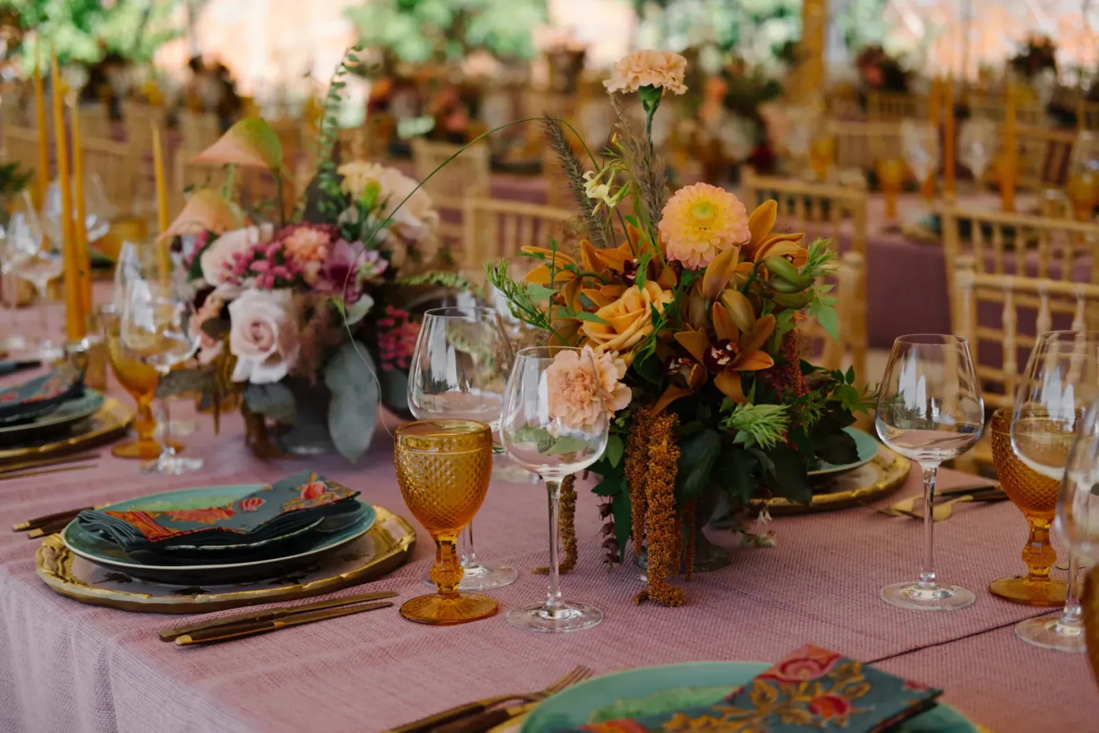 Elegant wedding table setting with pink tablecloth, gold-rimmed plates, amber glassware, and colorful floral centerpieces