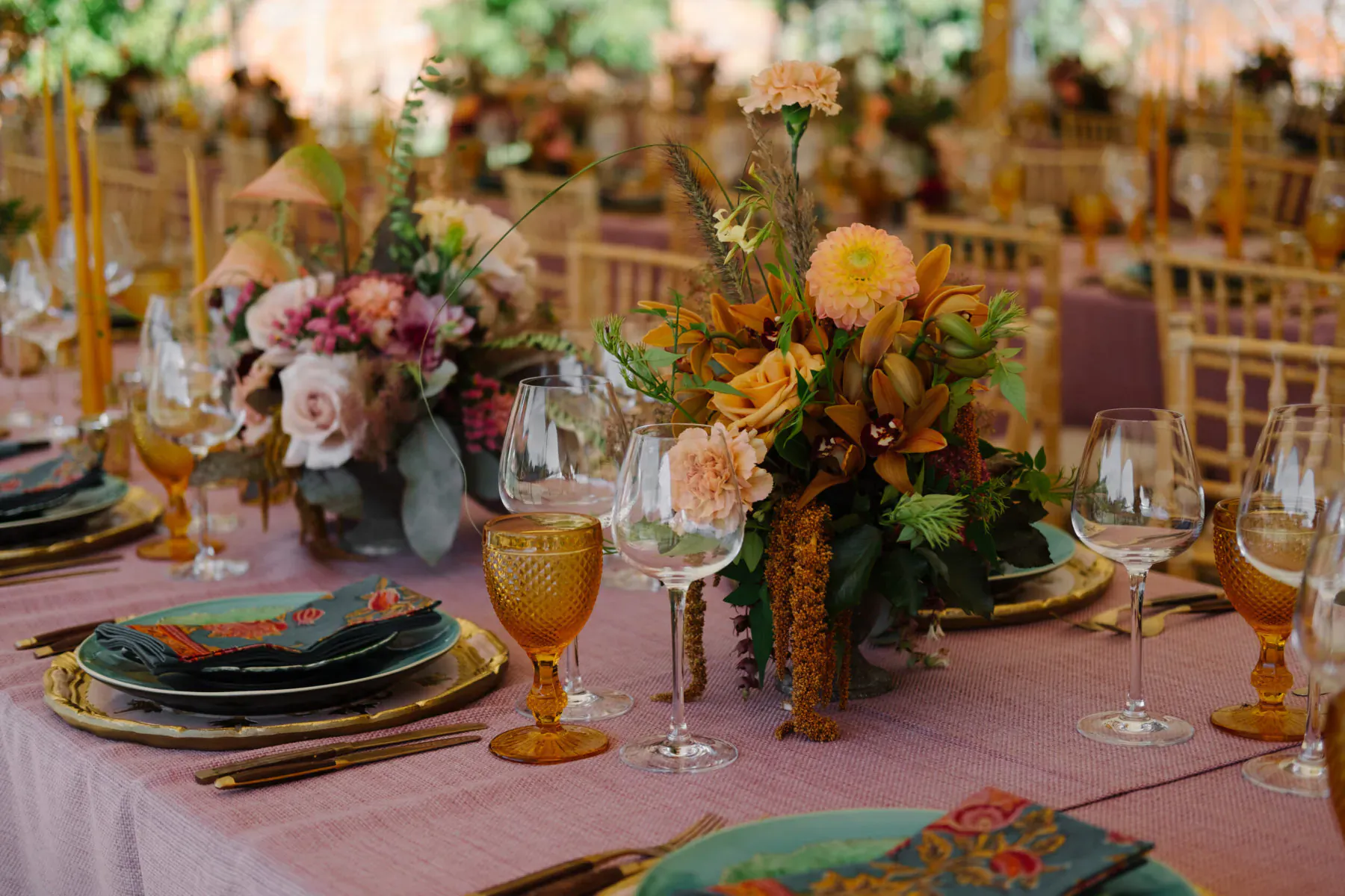 Elegant wedding table setting with pink tablecloth, gold-rimmed plates, amber glassware, and colorful floral centerpieces