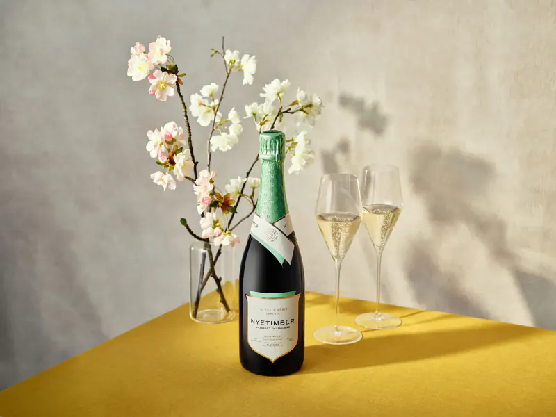 Nyetimber English sparkling wine bottle with filled flute glasses and cherry blossoms on yellow table for Mother's Day