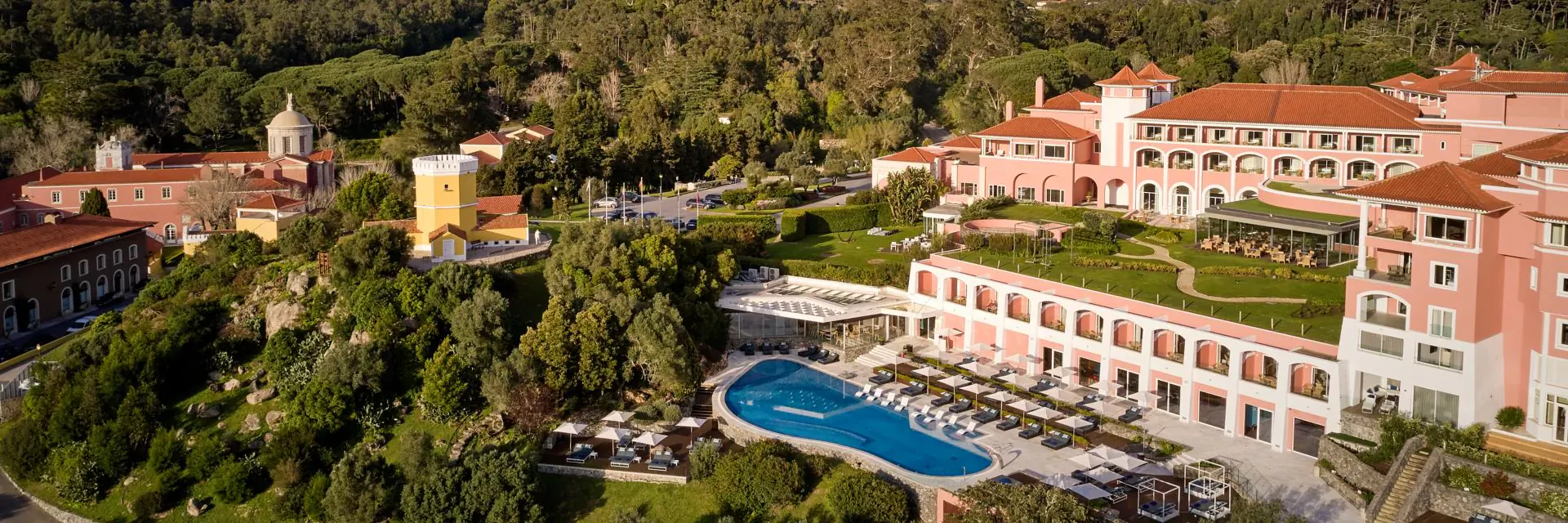 Aerial view of Penha Longa Resort in Portugal: pink buildings, blue pool, lush green gardens and hills.