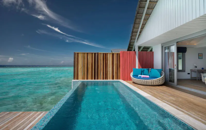 Cora Cora Maldives overwater villa with infinity pool overlooking turquoise lagoon, blue lounge chair on wooden deck.