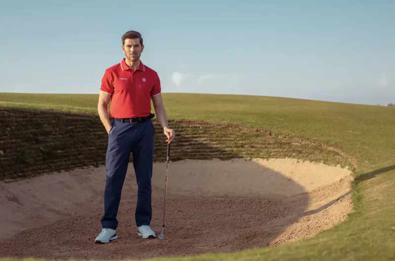 Man in red polo and navy pants stands by golf sand trap holding wedge on green course