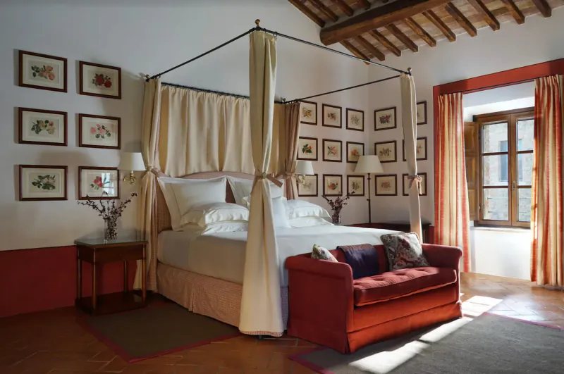 Elegant Tuscan bedroom with canopied bed, botanical art, red velvet bench, and arched windows.