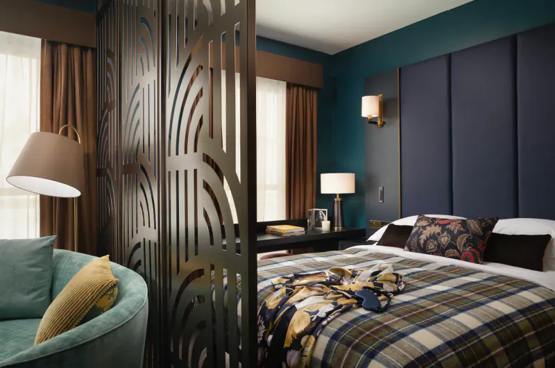 Elegant hotel room at The Other House South Kensington with teal walls, plaid bed, curved metal screen, and plush green armchair.