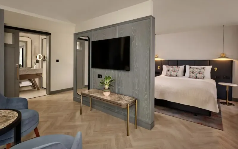 Modern hotel suite with king bed, TV on gray partition, en-suite bathroom, blue armchairs, and plants.
