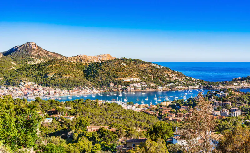 Aerial view of a picturesque Mediterranean harbor town with yachts, surrounded by green hills and mountains under blue sky.