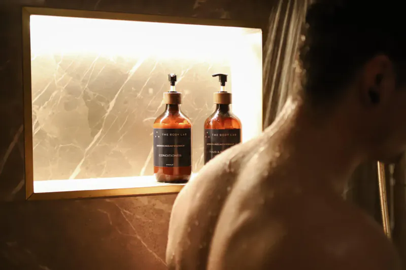 Man's wet back in shower, two amber pump bottles labeled 'The Body Lab' in frosted wall niche