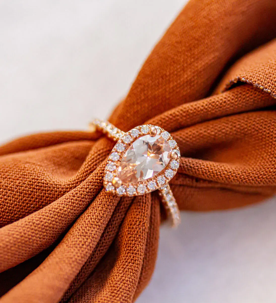 Pear-shaped rose gold diamond engagement ring with halo, displayed on terracotta fabric.