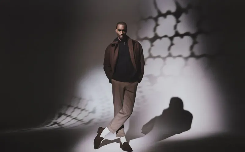 Man in brown cardigan and trousers stands confidently with crossed ankles against geometric light shadows, Falke gallery.