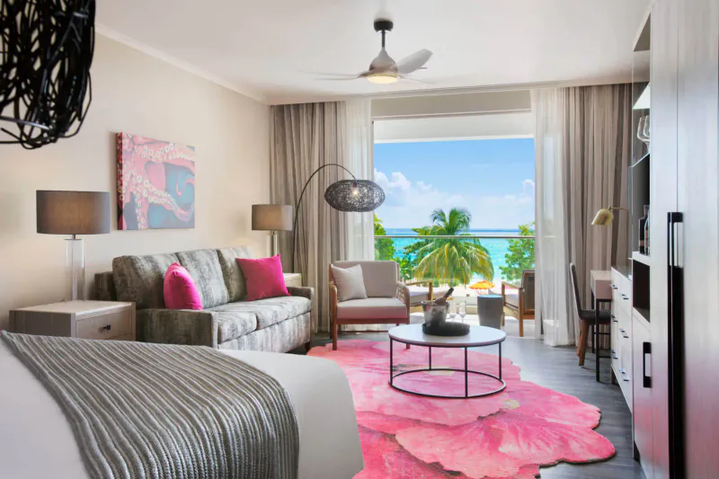 Luxurious O2 Beach Club suite in Barbados with pink rug, sofa, bed, and ocean-palm view balcony.