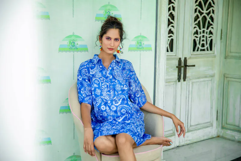 Woman in blue floral dress with green earrings sits on chair against green umbrella wallpaper and ornate white door