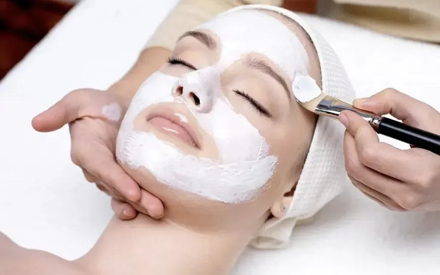 Close-up of esthetician applying white face mask to relaxed woman's face with brush in spa