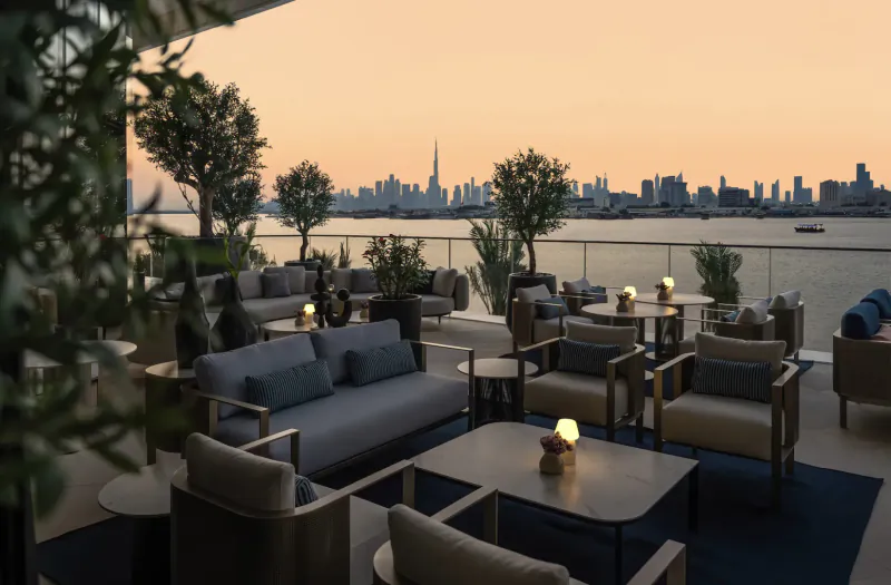 Evening rooftop lounge at Address Grand Creek Harbour with Dubai skyline, Burj Khalifa view, plush seating, plants, and lanterns.