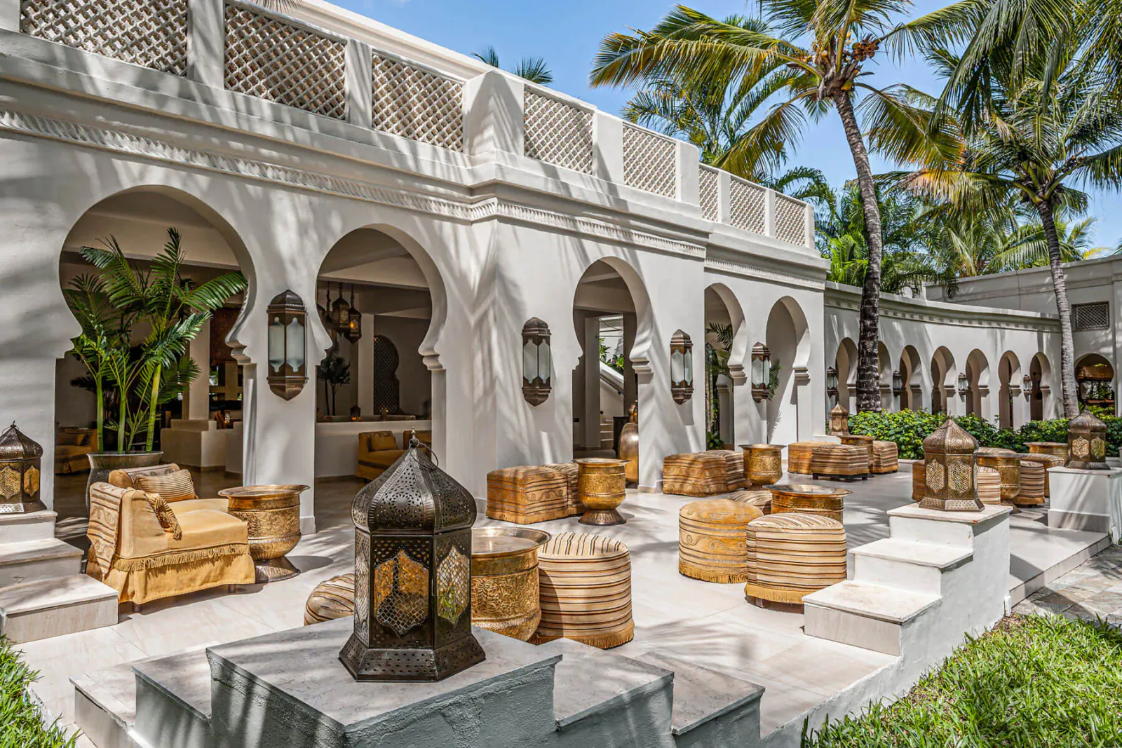 White arched Moroccan-style colonnade in Zanzibar resort courtyard with lanterns, wicker furniture, palms, and tropical plants under blue sky.