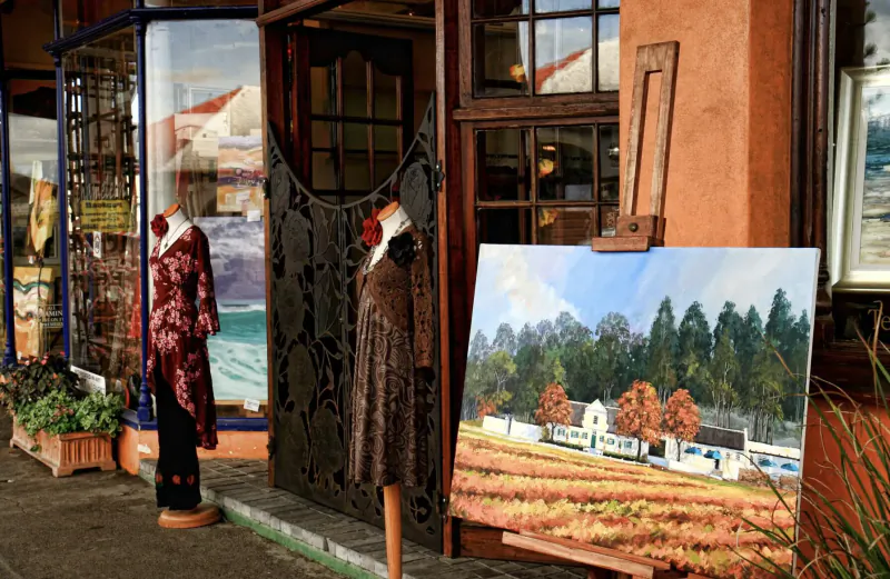Kalk Bay gallery exterior with mannequins in red and brown dresses, large landscape painting of farmhouse amid autumn trees on easel.