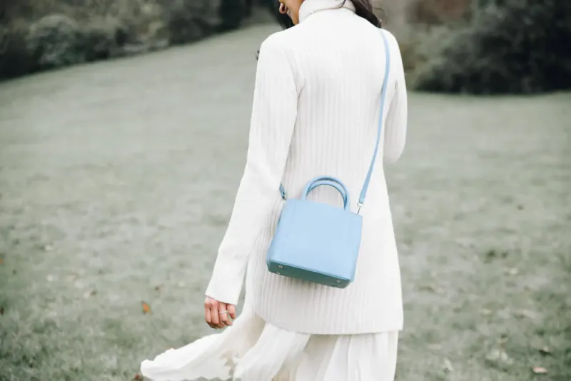 Woman in white turtleneck and skirt walks in green park, carrying light blue Luxtra handbag on shoulder