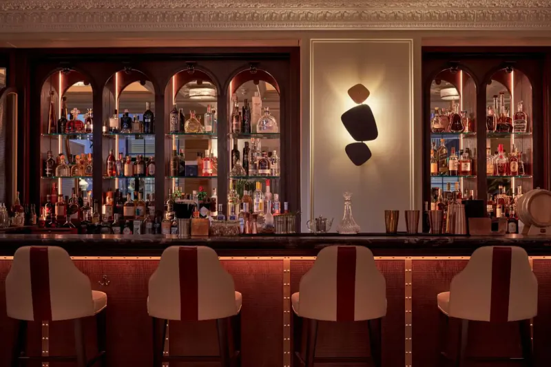Elegant hotel bar with arched wooden cabinets stocked with liquor bottles, modern wall art, and red-trimmed bar stools.