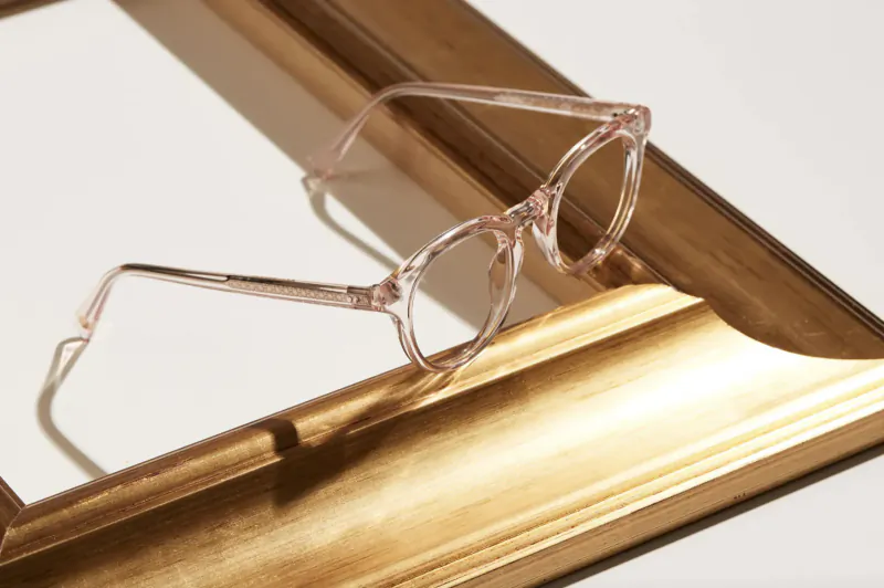 Pink transparent spectacles leaning against ornate gold picture frame, casting shadow.