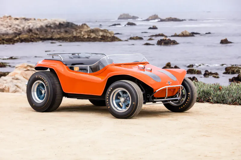 Bright orange dune buggy parked on sandy beach with ocean rocks and mist, Concours d'Elegance.