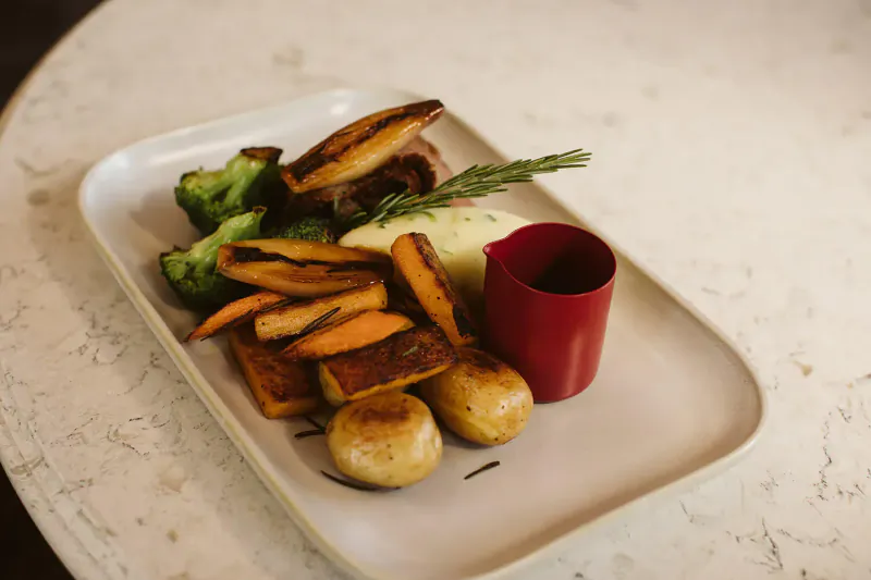 White plate with roasted beef, mashed potatoes, roasted carrots, broccoli, roast potatoes, rosemary, and red sauce cup on table.