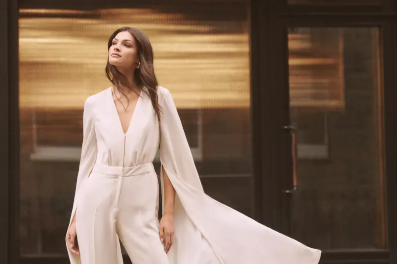 Woman in flowing white cape jumpsuit poses elegantly outside London boutique door on St. Christopher's Place.
