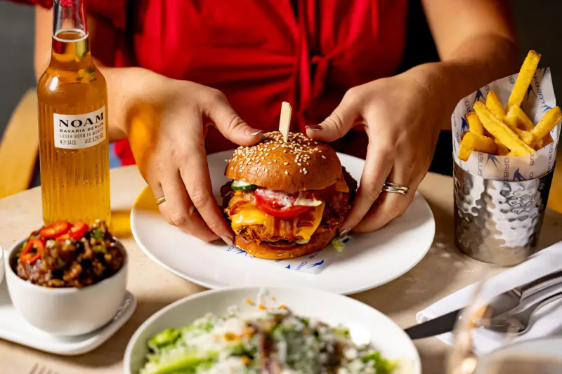 Woman in red top holding a Noam burger with fries, slaw, and spicy veg on plate beside beer bottle