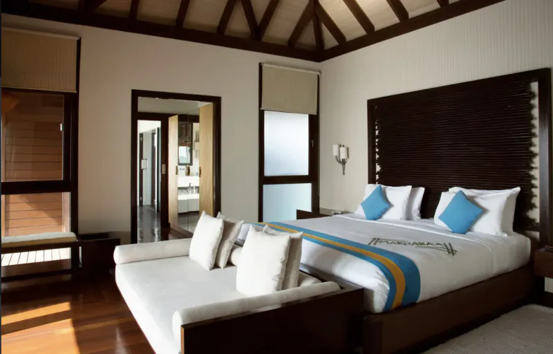 Luxurious bedroom in Coco Bodu Hithi resort with king bed, white sofa, wooden accents, and ocean-view doors.