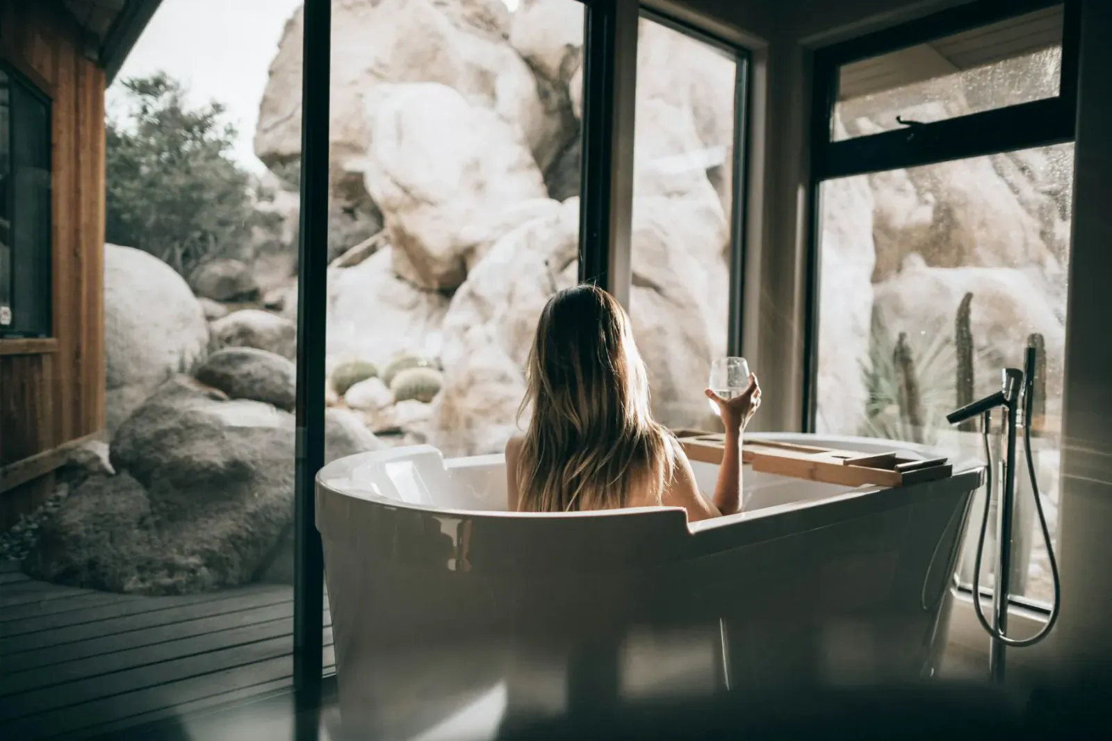Woman with long blonde hair relaxes nude in white bathtub, holding wine glass, scenic rocky mountain view through window